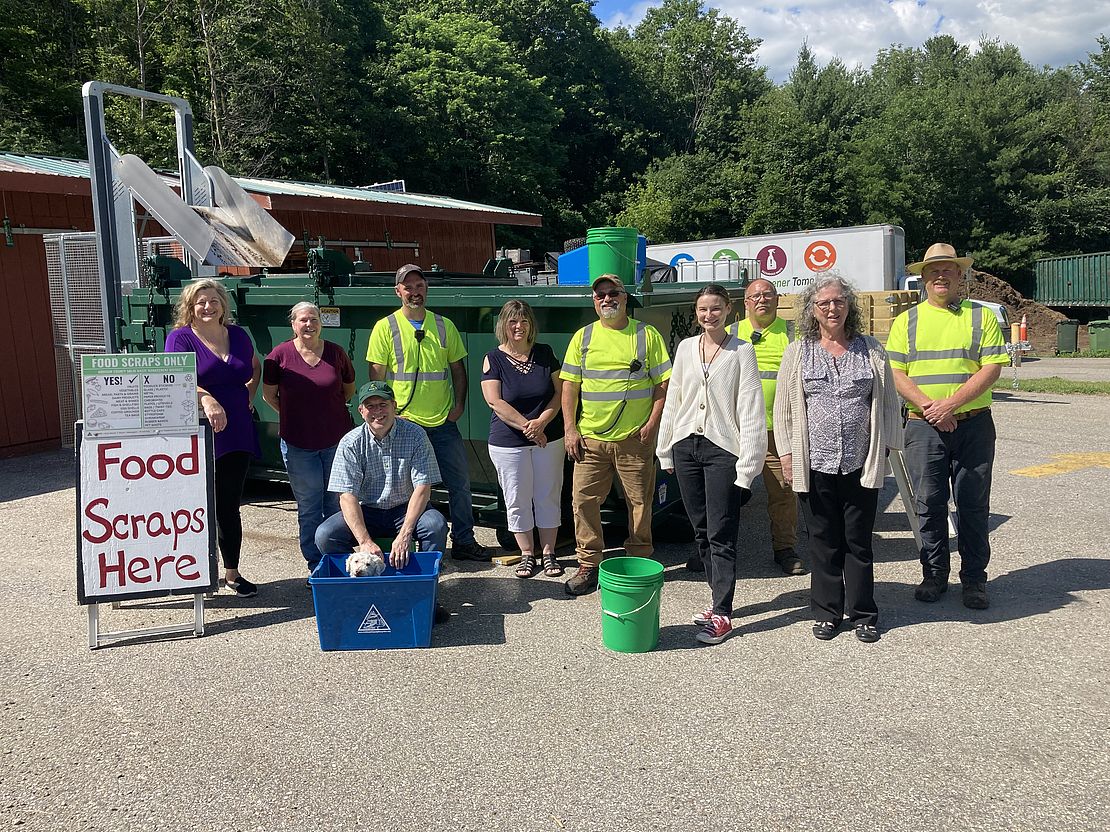 A photo of the staff of ACSWMD: Patti Johnson, Shelly Edson, Chad Kimball, Chantel Bolduc, Gene Pouliot, Brian Sherwin, Gary Hobbs, Don Maglienti, Emily Johnston, and Teri Kuczynski stand shoulder to shoulder smiling. Pepper the dog sits in a recycling bin.